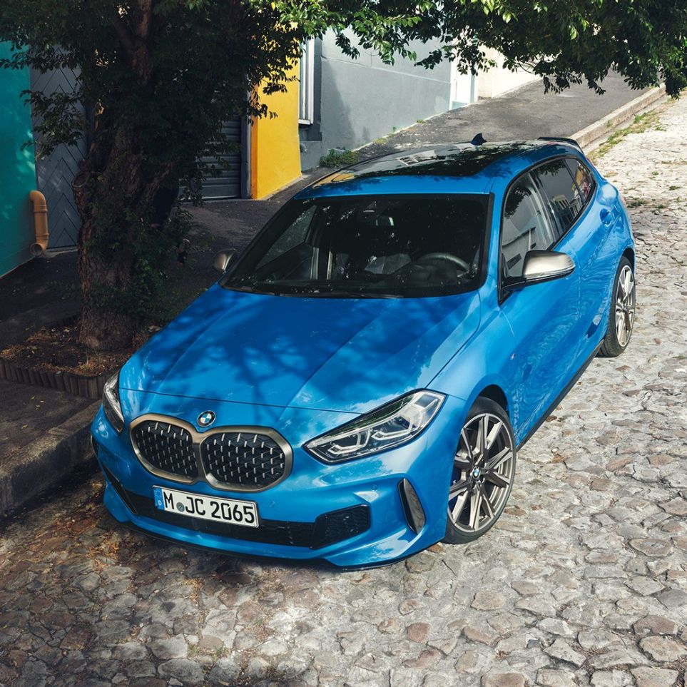 1 serijos BMW šalikelėje Misano Blue mėlyno metaliko spalvos BMW 1 serijos modelis, trijų ketvirčių vaizdas iš priekio, automobilis parkuojamas lygiagrečiai kelio, vaizdas iš paukščio skrydžio