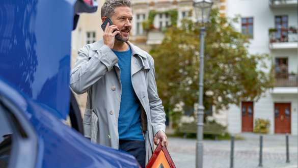 Vyras stovi prie BMW bagažinės Vyras stovi prie BMW bagažinės su išmaniuoju telefonu ir įspėjamuoju trikampiu ženklu rankose