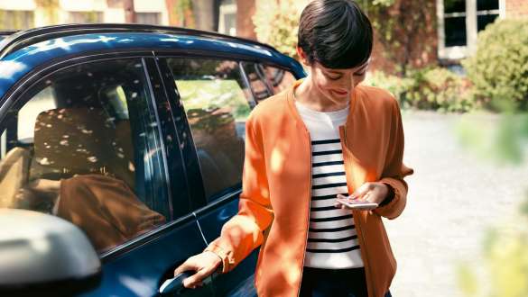Moteris žiūri į savo išmanųjį telefoną ir atidaro savojo BMW dureles BMW telepaslaugos, moteris žiūri į savo išmanųjį telefoną ir atidaro savojo BMW dureles