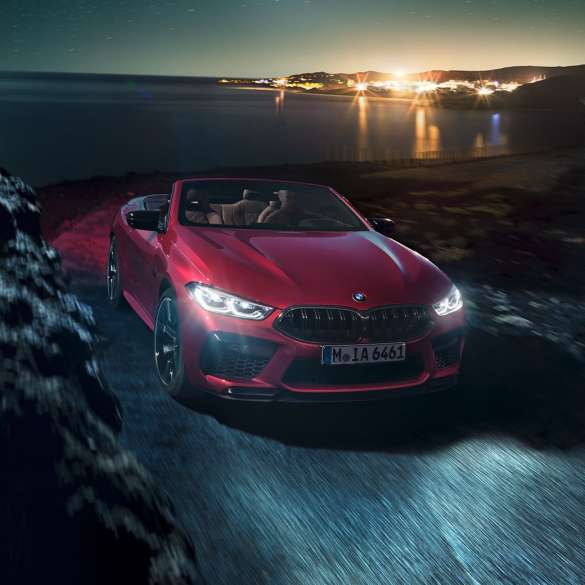 BMW M8 Competition kabrioletas, trijų ketvirčių vaizdas iš priekio. BMW M8 Competition kabrioletas Motegi Red raudono metaliko spalvos, su atviru minkštu stogu trijų ketvirčių naktinis važiuojančio pakrante automobilio vaizdas iš priekio.