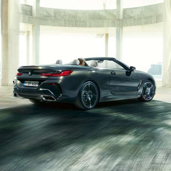 BMW M850i xDrive kabrioletas, trijų ketvirčių vaizdas iš galo. BMW M850i xDrive kabrioletas Dravit Grey pilko metaliko spalvos su atviru minkštu stogu, trijų ketvirčių pro pastatą važiuojančio automobilio vaizdas iš galo.