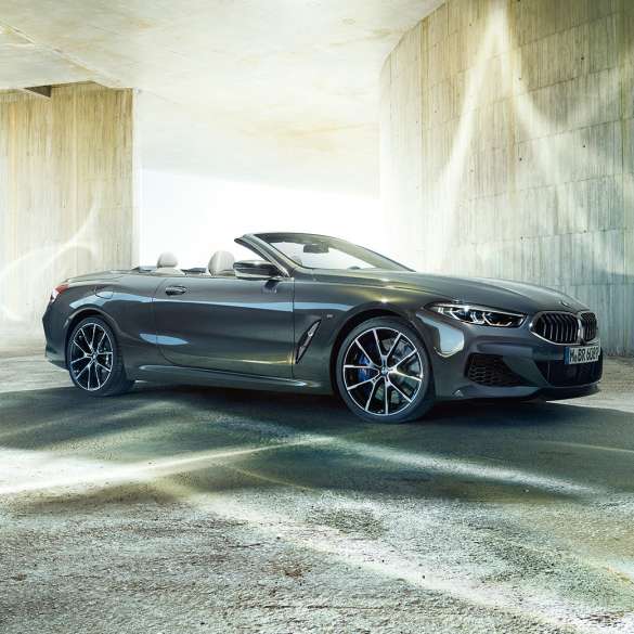 BMW M850i xDrive kabrioletas, trijų ketvirčių vaizdas iš šono. BMW M850i xDrive kabrioletas Dravit Grey pilko metaliko spalvos su atviru stogu, trijų ketvirčių priešais pastatą stovinčio automobilio vaizdas iš šono.