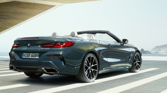 BMW M850i xDrive kabrioletas, trijų ketvirčių vaizdas iš galo. BMW M850i xDrive kabrioletas Dravit Grey pilko metaliko spalvos su atviru minkštu stogu, trijų ketvirčių parkuojamo automobilio vaizdas iš galo vandenyno fone.