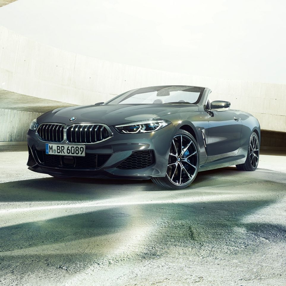 BMW M850i xDrive kabrioletas, trijų ketvirčių vaizdas iš priekio. BMW M850i xDrive kabrioletas Dravit Grey pilko metaliko spalvos, trijų ketvirčių vaizdas iš priekio, stovintis priešais betoninį pastatą.