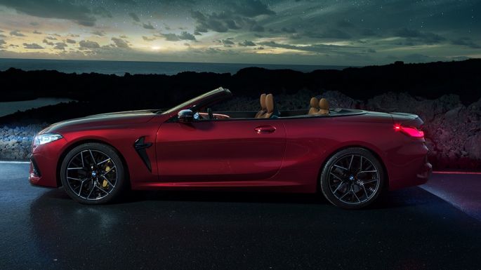 BMW M8 Competition kabrioletas, vaizdas iš šono. BMW M8 Competition kabrioletas Motegi Red raudono metaliko spalvos su atviru minkštu stogu, automobilio, stovinčio šalia vyro žvaigždėto dangaus fone, vaizdas iš šono.