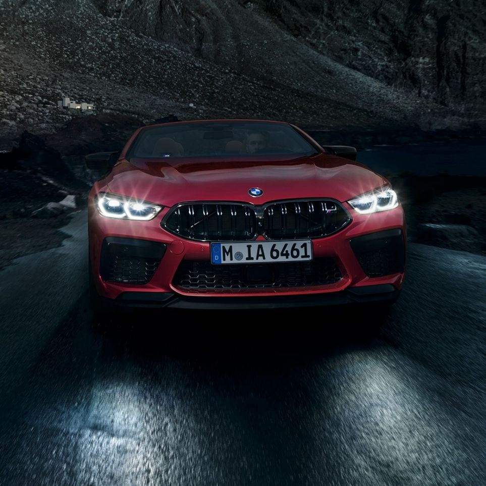 BMW M8 Competition kabrioletas, vaizdas iš priekio. BMW M8 Competition kabrioletas Motegi Red raudono metaliko spalvos, su šviečiančiais priekiniais žibintais, važiuojančio naktį automobilio vaizdas iš priekio.