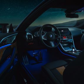 BMW M8 Competition kupė, interjeras BMW M8 Competition kupė, interjero su M odiniu vairu ir mėlynu aplinkos apšvietimu bei atviromis vairuotojo durelėmis vaizdas naktį