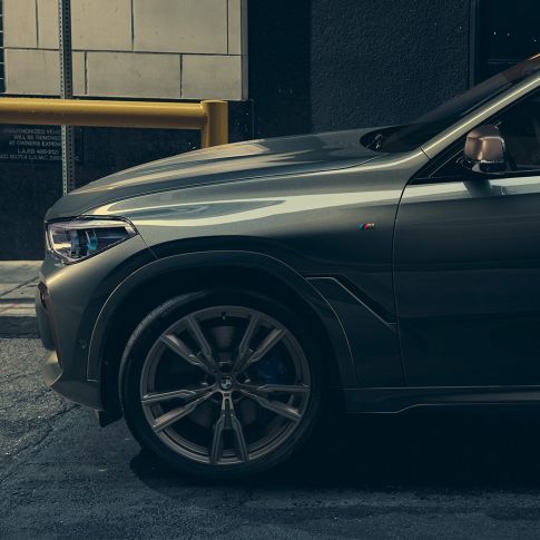 BMW X6 M50i vaizdas iš šono Vyras, atsirėmęs į BMW X6 M50i miesto fone, vaizdas iš šono