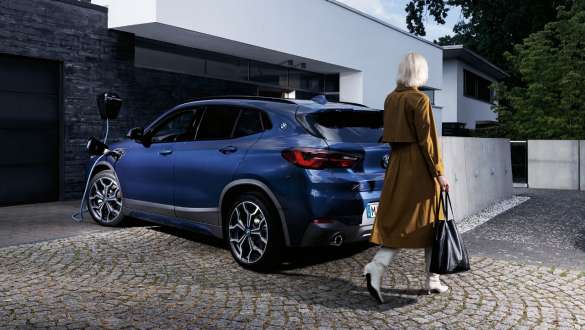 Įkraunamas hibridinis BMW X2 xDrive25e (F39), įkraunamo automobilio trijų ketvirčių vaizdas iš galo Įkraunamas hibridinis BMW X2 xDrive25e (F39), trijų ketvirčių vaizdas iš galo su modeliu įkrovimo stotelėje