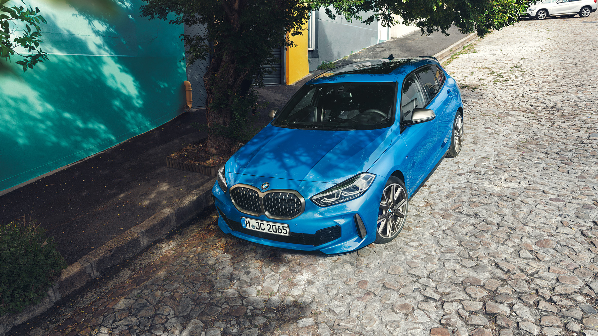 1 serijos BMW šalikelėje Misano Blue mėlyno metaliko spalvos BMW 1 serijos modelis, trijų ketvirčių vaizdas iš priekio, automobilis parkuojamas lygiagrečiai kelio, vaizdas iš paukščio skrydžio