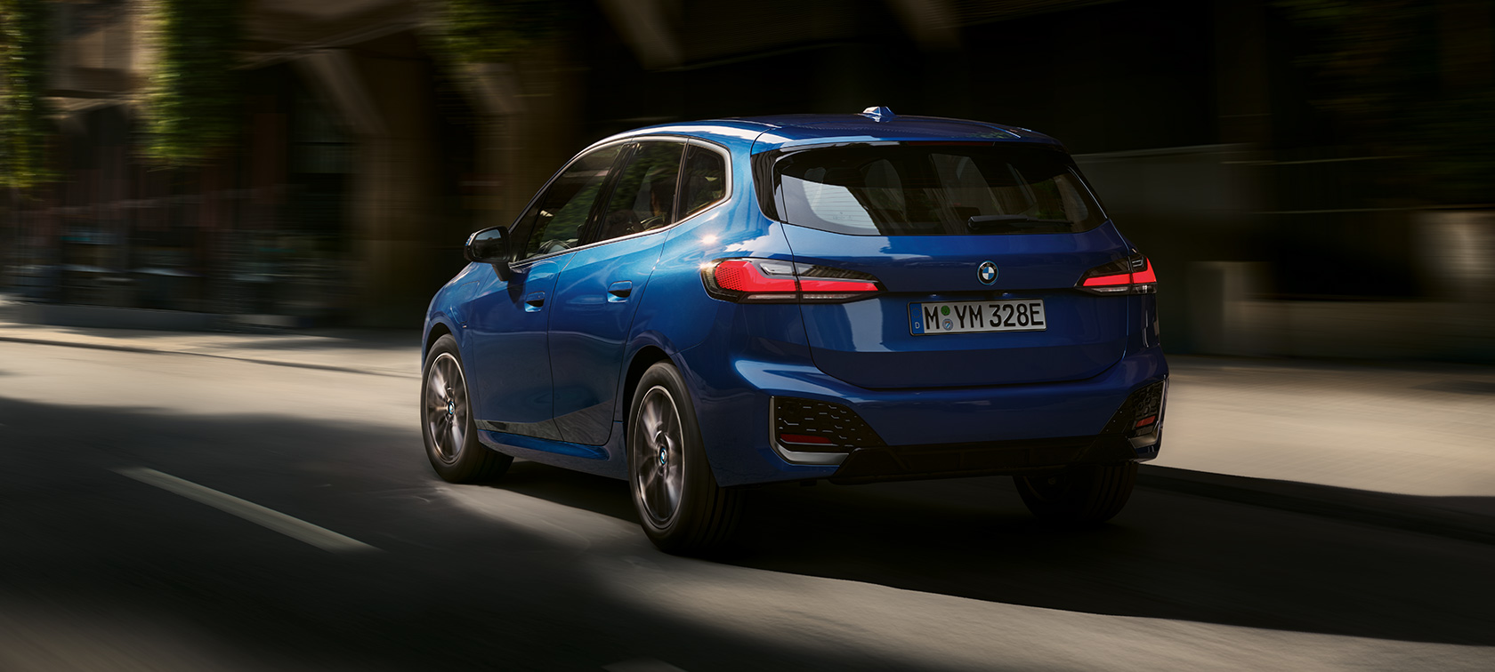 BMW 2 serijos Active Tourer 230e įkraunamas hibridas (U06) 2021, mėlynos M Portimao Blue metalizuotos spalvos, važiuojančio automobilio trijų ketvirčių vaizdas iš galo miesto fone