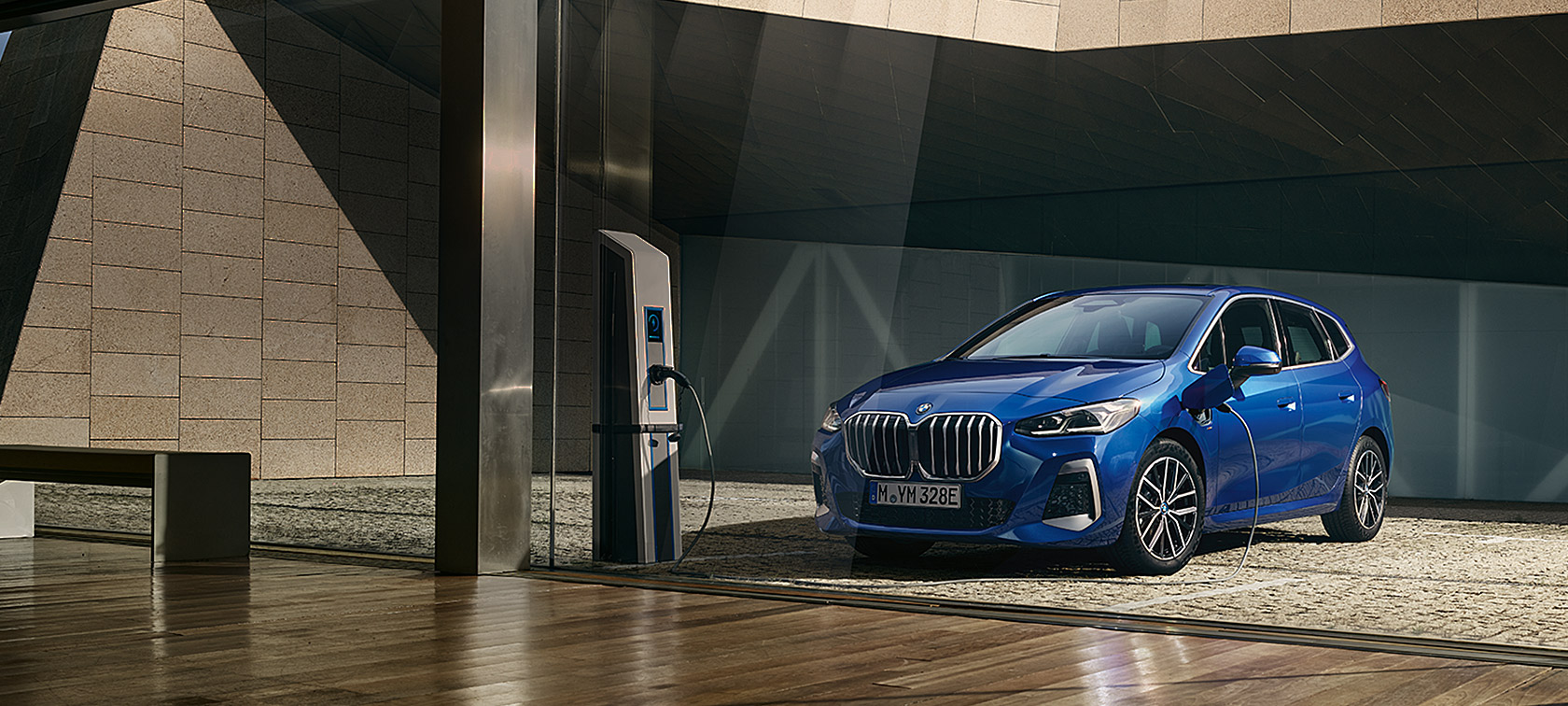 BMW 2 serijos Active Tourer 230e įkraunamas hibridas (U06) 2021, mėlynos M Portimao Blue metalizuotos spalvos, trijų ketvirčių vaizdas iš priekio prie įkrovimo stotelės