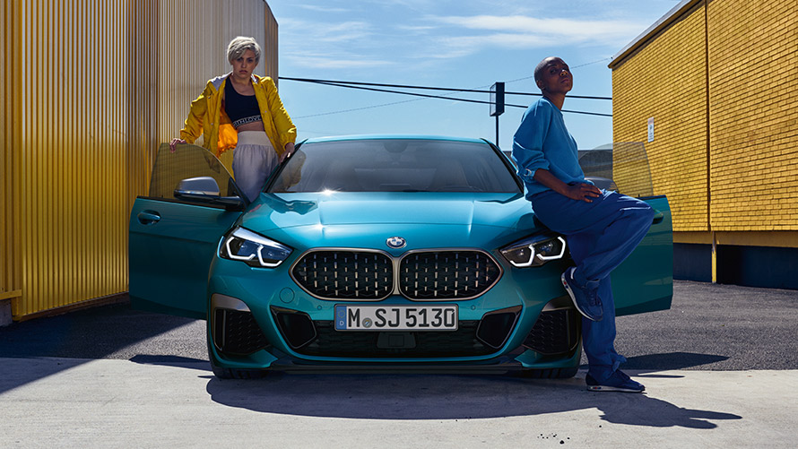BMW 2 serijos Gran kupė (F44), vaizdas iš priekio BMW 2 serijos Gran kupė (F44) mėlynos metalizuotos <i>Snapper Rocks Blue</i> kėbulo spalvos, vaizdas iš priekio su atviromis durelėmis ir dviem žmonėmis automobilyje