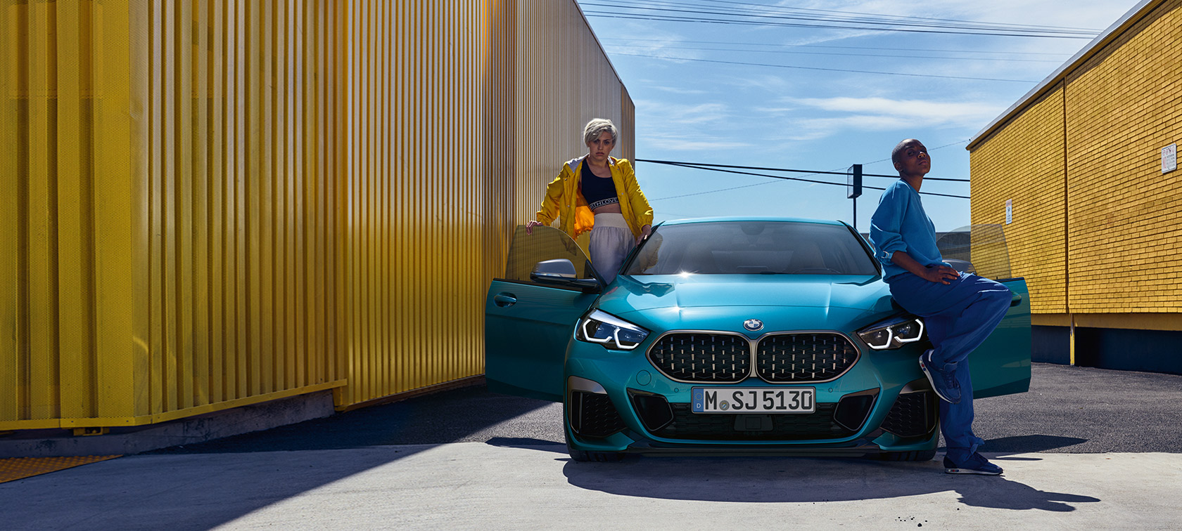 BMW 2 serijos Gran kupė (F44), vaizdas iš priekio BMW 2 serijos Gran kupė (F44) mėlynos metalizuotos <i>Snapper Rocks Blue</i> kėbulo spalvos, vaizdas iš priekio su atviromis durelėmis ir dviem žmonėmis automobilyje