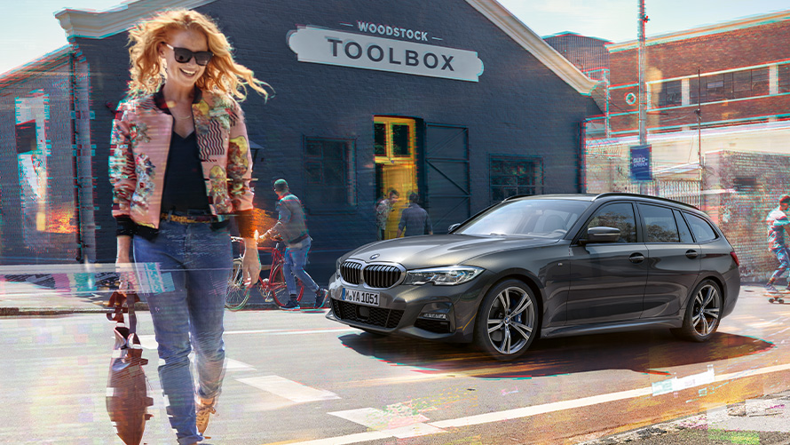 BMW 3 serijos Touring (G21) trijų ketvirčių vaizdas iš priekio su moterimi medienos darbams skirtų įrankių parduotuvės fone Gatvę kertanti moteris priešais BMW 3 serijos Touring, (G21) medienos darbams skirtų įrankių parduotuvės fone