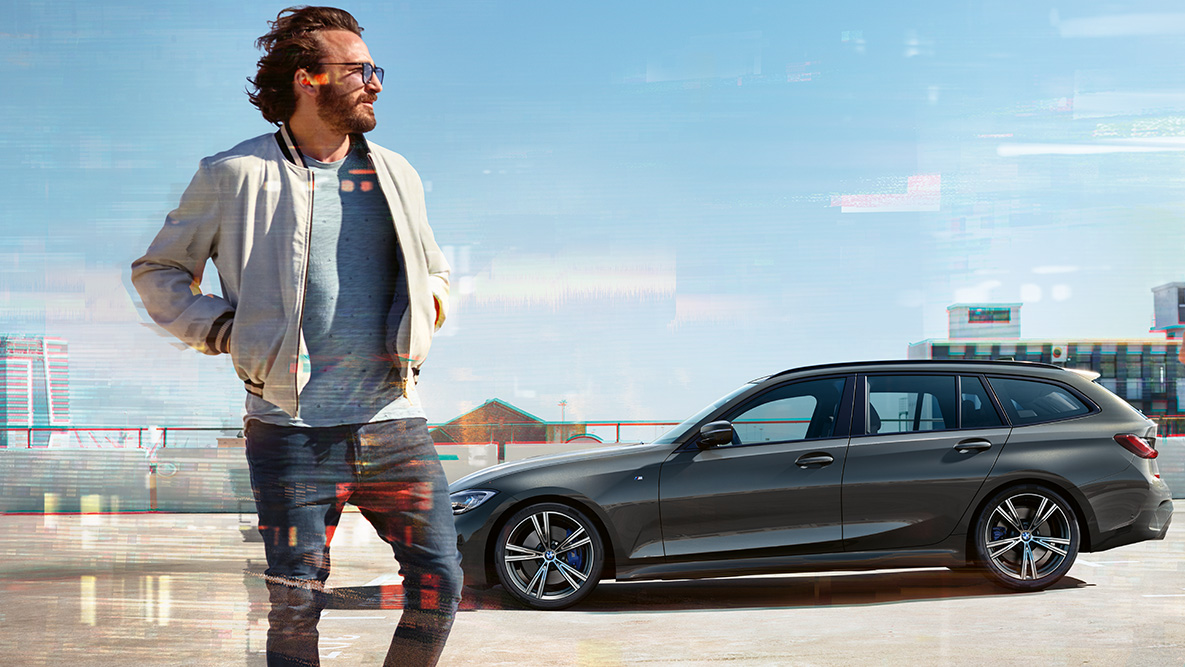 BMW 3 serijos Touring, (G21), trijų ketvirčių vaizdas iš priekio BMW 3 serijos Touring (G21), pilkos <i>Dravite Grey</i> spalvos, užmiesčio fone ant tilto važiuojančio automobilio trijų ketvirčių vaizdas iš priekio