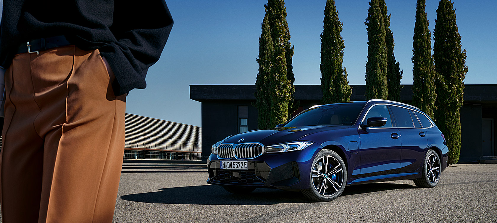 BMW 3 serijos Touring LCI G21 mėlynos Tansanit Blue II spalvos, medžių fone stovinčio automobilio trijų ketvirčių vaizdas iš priekio