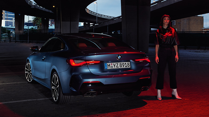 BMW 4 serijos kupė (G22) 2020 mėlynos metalizuotos Artic Race Blue spalvos, vaizdas iš galo su žmogumi šalia BMW 4 serijos kupė (G22) 2020 mėlynos metalizuotos Artic Race Blue spalvos, vaizdas iš galo su žmogumi šalia