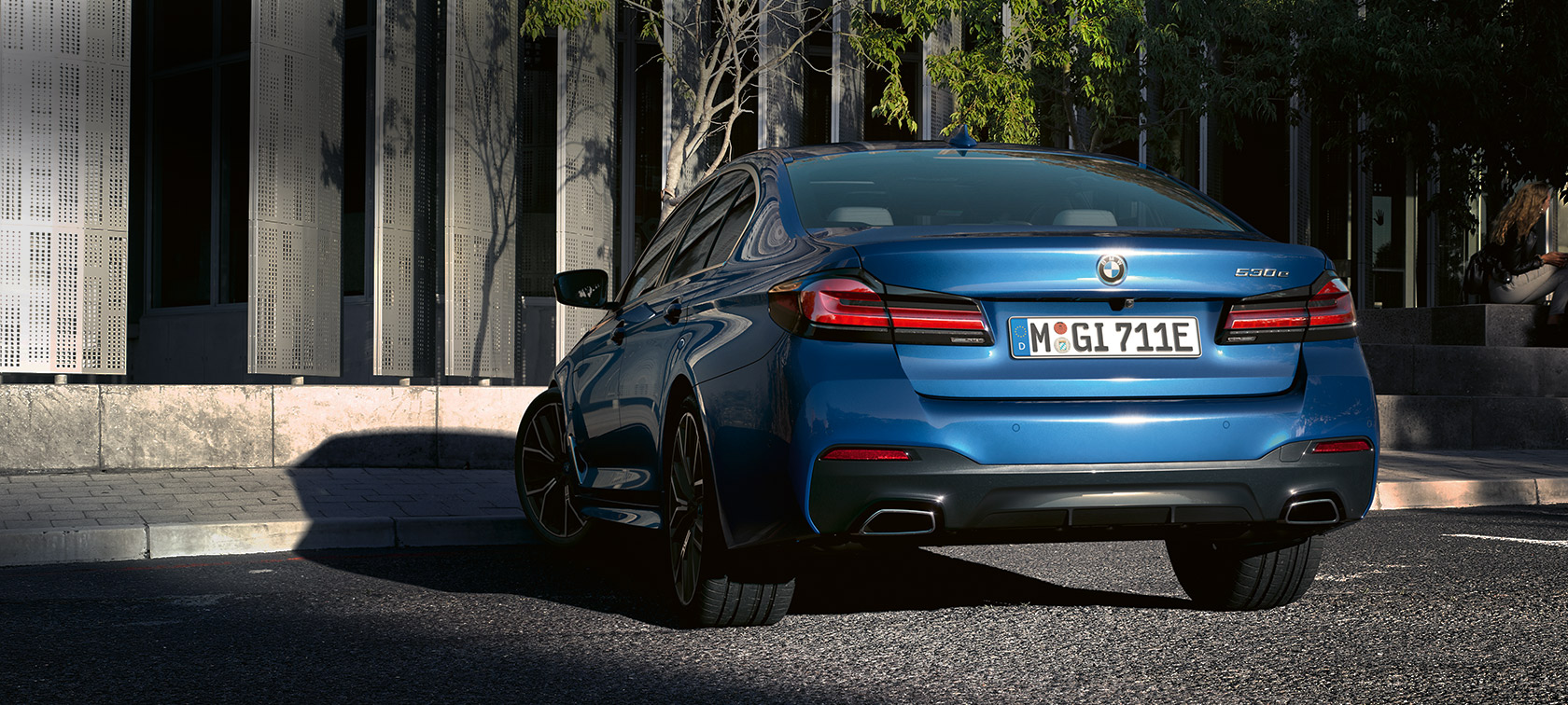 BMW 5 serijos sedanas eDrive (G30), mėlynos Phytonic Blue metalizuotos spalvos BMW 5 serijos sedanas eDrive (G30), mėlynos Phytonic Blue metalizuotos spalvos, pastato fone parkuojamo automobilio vaizdas iš galo