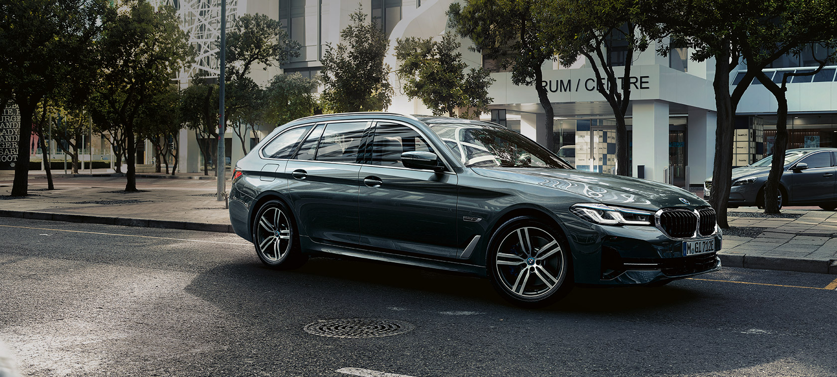 BMW 5 serijos Touring (G31) Facelift 2020 pilkos metalizuotos Sophisto Grey spalvos,važiuojančio automobilio 7/8 vaizdas iš priekio miesto pakraštyje