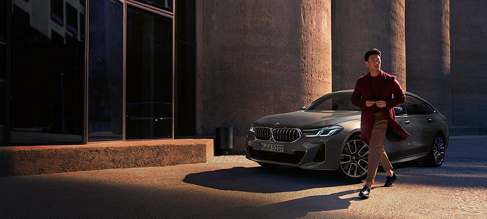 BMW 6 serijos Gran Turismo 640i xDrive (G32) 2020 pilkos Bernina Grey spalvos, trijų ketvirčių vaizdas iš priekio su šalia stovinčiu vyru