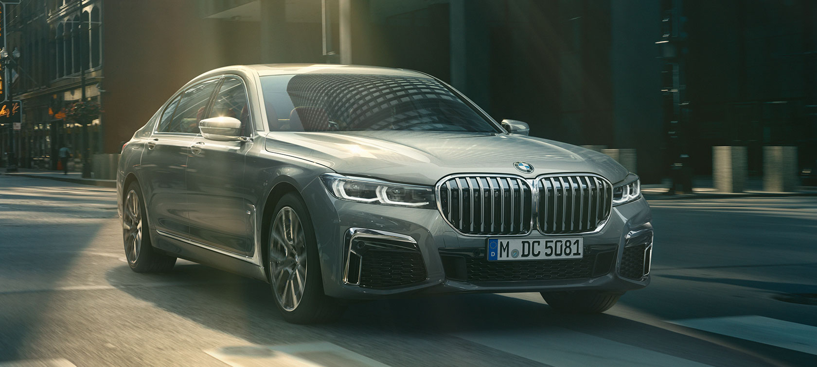 Keliu važiuojantis pilkas BMW 7 serijos sedanas, trijų ketvirčių vaizdas iš priekio Miesto fone važiuojantis pilkas BMW 7 serijos sedanas, trijų ketvirčių vaizdas iš priekio