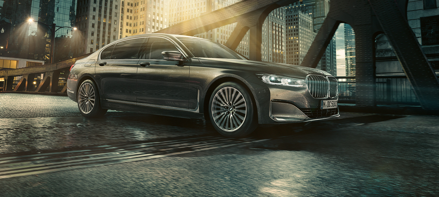BMW 7 serijos sedano (G11 G12) 2019 vaizdas iš šono BMW 7 serijos sedanas (G11 G12) 2019 pilkos Bernina Grey spalvos su tviskančio gintaro efektu, miesto fone važiuojančio automobilio trijų ketvirčių vaizdas iš šono