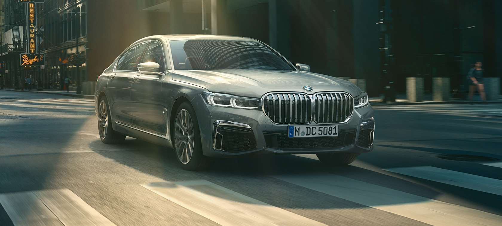 BMW 7 serijos sedanas (G11 G12) 2019 pilkos Bernina Grey spalvos su tviskančio gintaro efektu, trijų ketvirčių vaizdas iš priekio BMW 7 serijos sedanas (G11 G12) 2019 pilkos Bernina Grey spalvos su tviskančio gintaro efektu, miesto fone važiuojančio automobilio trijų ketvirčių vaizdas iš priekio