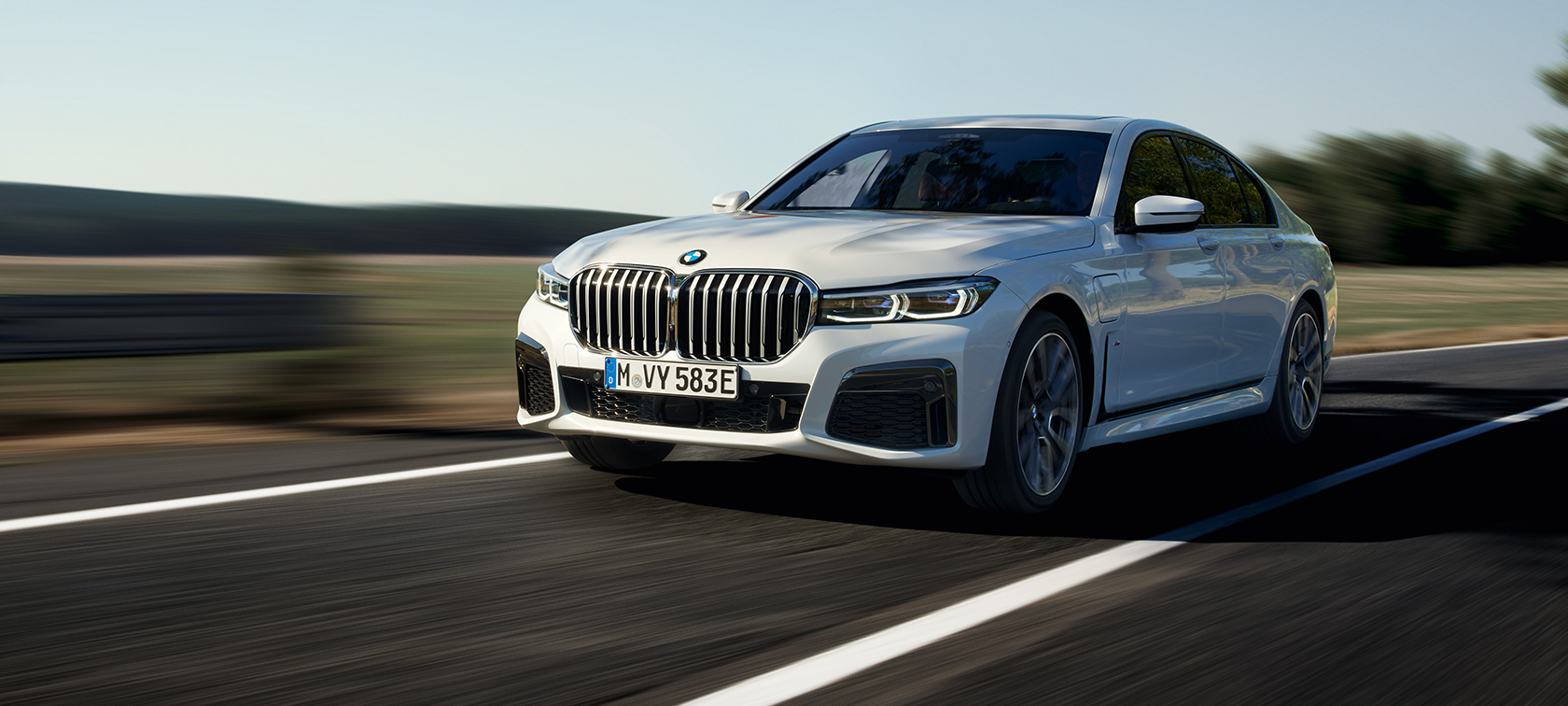 BMW 745e įkraunamas hibridinis sedanas (G11 G12) baltos metalizuotos Mineral White kėbulo spalvos, trijų ketvirčių vaizdas iš priekio BMW 745e įkraunamas hibridinis sedanas (G11 G12) baltos metalizuotos Mineral White kėbulo spalvos, užmiesčio keliu važiuojančio automobilio trijų ketvirčių vaizdas iš priekio