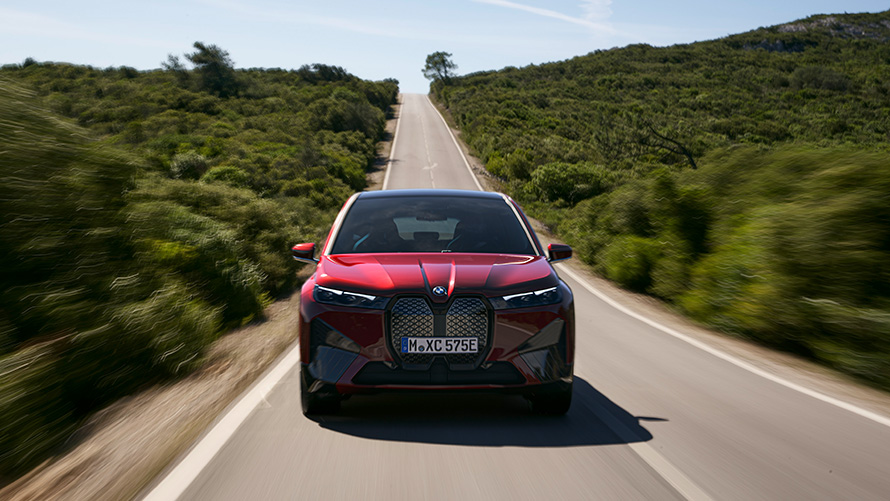 BMW iX i20 elektrinis SUV BMW iX xDrive50, nuvažiuojamas atstumas 2021 m. elektrinis padidinto pravažumo BMW iX i20 automobilis, gatve važiuojantis raudonos Aventurin Red spalvos BMW iX xDrive50 automobilis, nuvažiuojamas atstumas