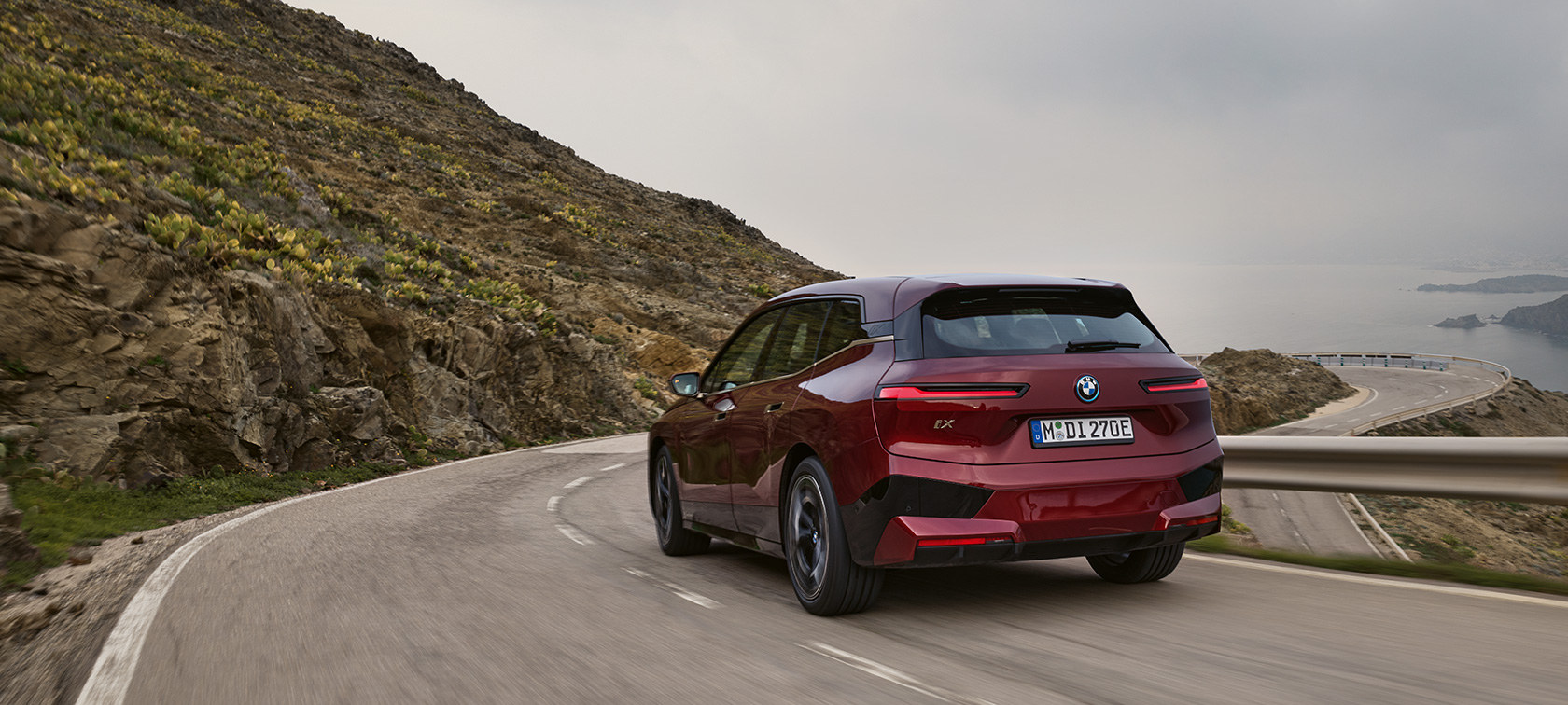 BMW iX (i20) 2021 elektrinis SUV BMW iX xDrive50, pakrantės keliu važiuojančio automobilio vaizdas iš galo BMW iX (i20) 2021 elektrinis SUV BMW iX xDrive50 raudonos Aventurin Red spalvos, pakrantės keliu važiuojančio automobilio vaizdas iš galo