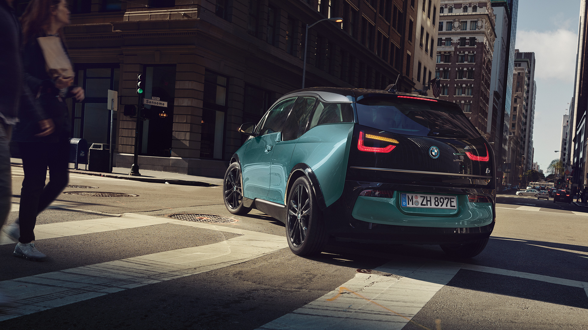 BMW i3 2018 važiuojančio automobilio trijų ketvirčių vaizdas iš galo BMW i3 I01 2018 Jucaro beige smėlio spalvos su Frozen Grey pilko metaliko spalvos akcentu, miesto fone važiuojančio automobilio trijų ketvirčių vaizdas iš galo