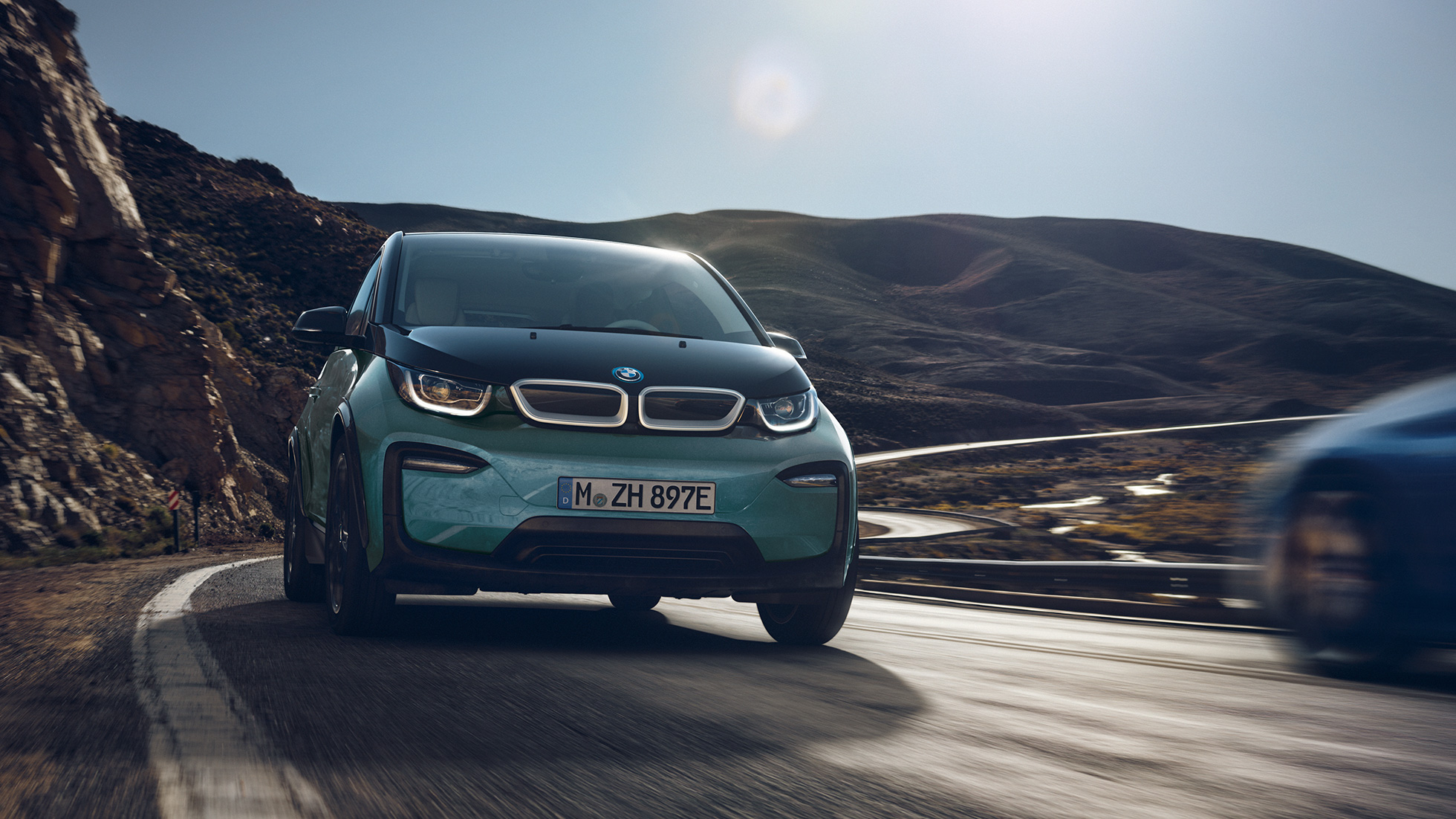 BMW i3 2018 važiuojančio automobilio vaizdas iš šono BMW i3 I01 2018 Jucaro beige smėlio spalvos su Frozen Grey pilko metaliko spalvos akcentu, miesto fone važiuojančio automobilio vaizdas iš šono