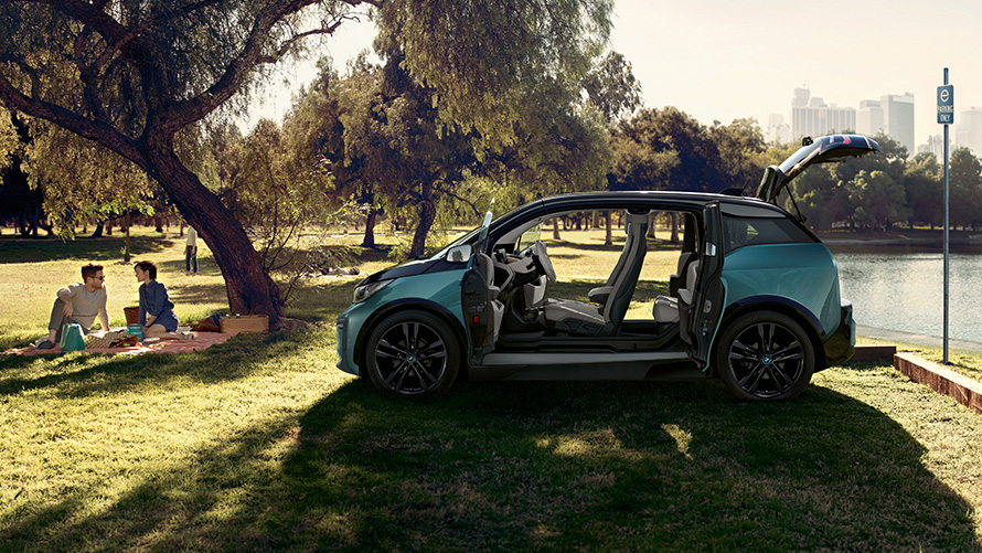 BMW i3 2018 parke BMW i3 I01 2018 Jucaro beige smėlio spalvos su Frozen Grey pilko metaliko spalvos akcentu, vaizdas iš šono su atviromis durelėmis parke