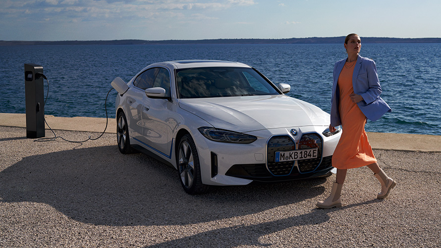 BMW i4 eDrive40 (G26) 2021 trijų ketvirčių vaizdas iš priekio įkrovimo stotelėje BMW i4 eDrive40 (G26) 2021 baltos Mineral White spalvos, trijų ketvirčių vaizdas iš priekio įkrovimo stotelėje prie ežero