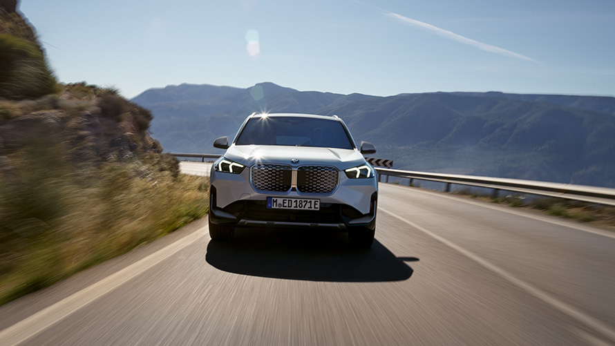 BMW iX1 U11 BEV 2022 vaizdas iš priekio BMW iX1 U11 BEV 2022 baltos metalizuotos Mineral White spalvos, kalnų keliu važiuojančio automobilio vaizdas iš priekio
