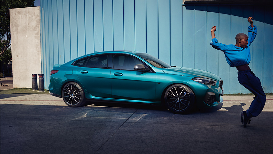 BMW M235i xDrive Gran kupė (F44) vaizdas iš šono su moterimi šalia BMW M235i xDrive Gran kupė (F44) mėlynos metalizuotos <i>Snapper Rocks Blue</i> spalvos, vaizdas iš šono su moterimi šalia