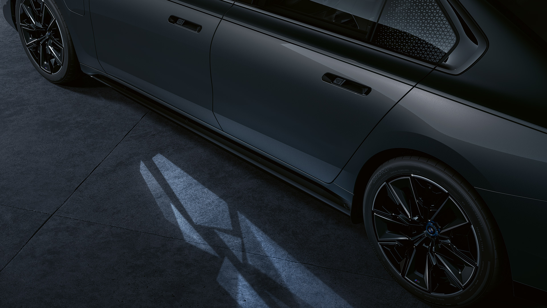 Apšvietimo koncepcija BMW M760e xDrive sedanas G70 Apšvietimo koncepcija BMW M760e xDrive sedanas G70 BMW Individual dviejų atspalvių Black Sapphire juoda metalizuota dažų apdaila BMW Individual Dravite Grey pilka metalizuota pilka spalva priartintas vaizdas iš šono su pasitikimo apšvietimu Welcome Light Carpet