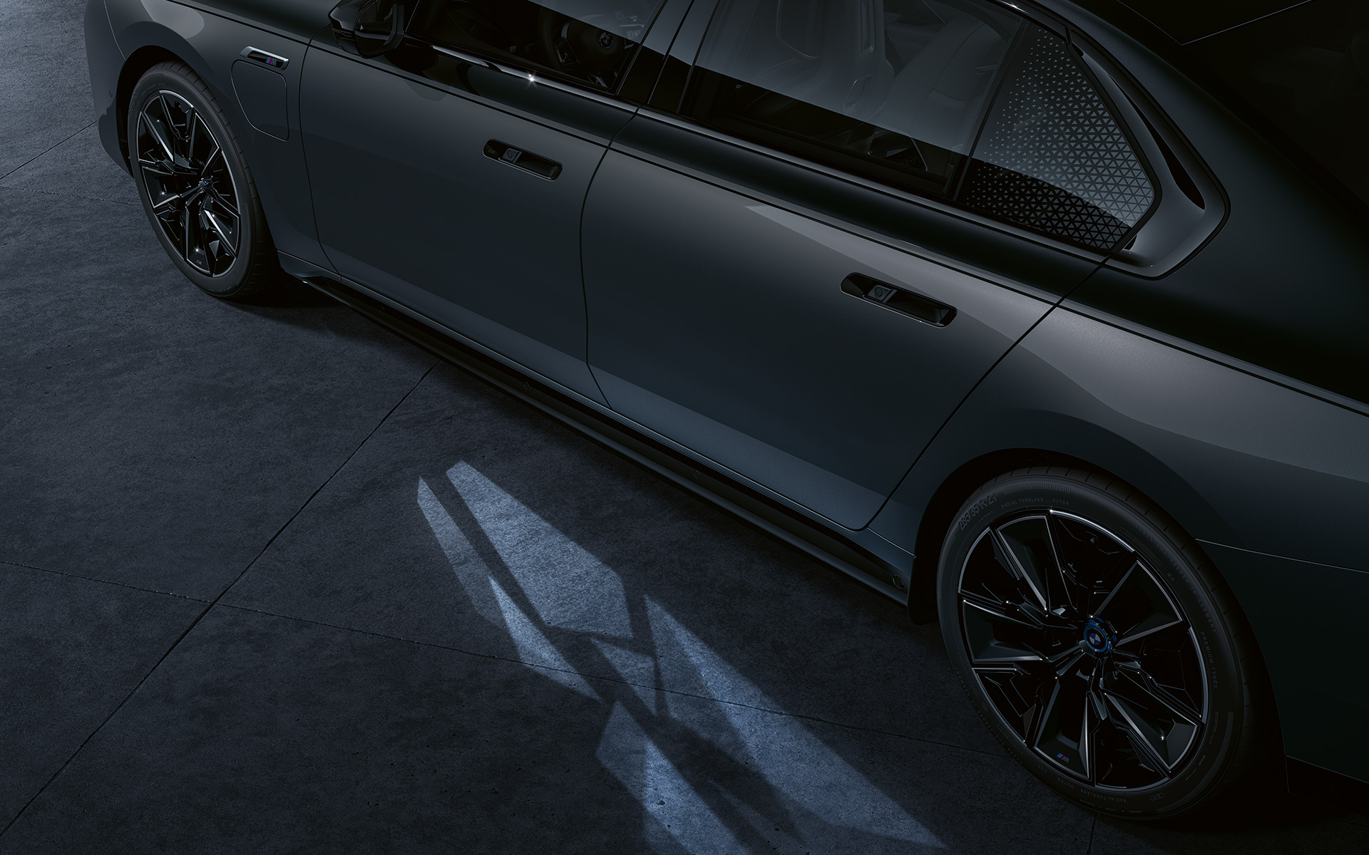 Apšvietimo koncepcija BMW M760e xDrive sedanas G70 BMW Individual dviejų atspalvių Black Sapphire juoda metalizuota dažų apdaila BMW Individual Dravite Grey pilka metalizuota spalva priartintas vaizdas iš šono su pasitikimo apšvietimu Welcome Light Carpet