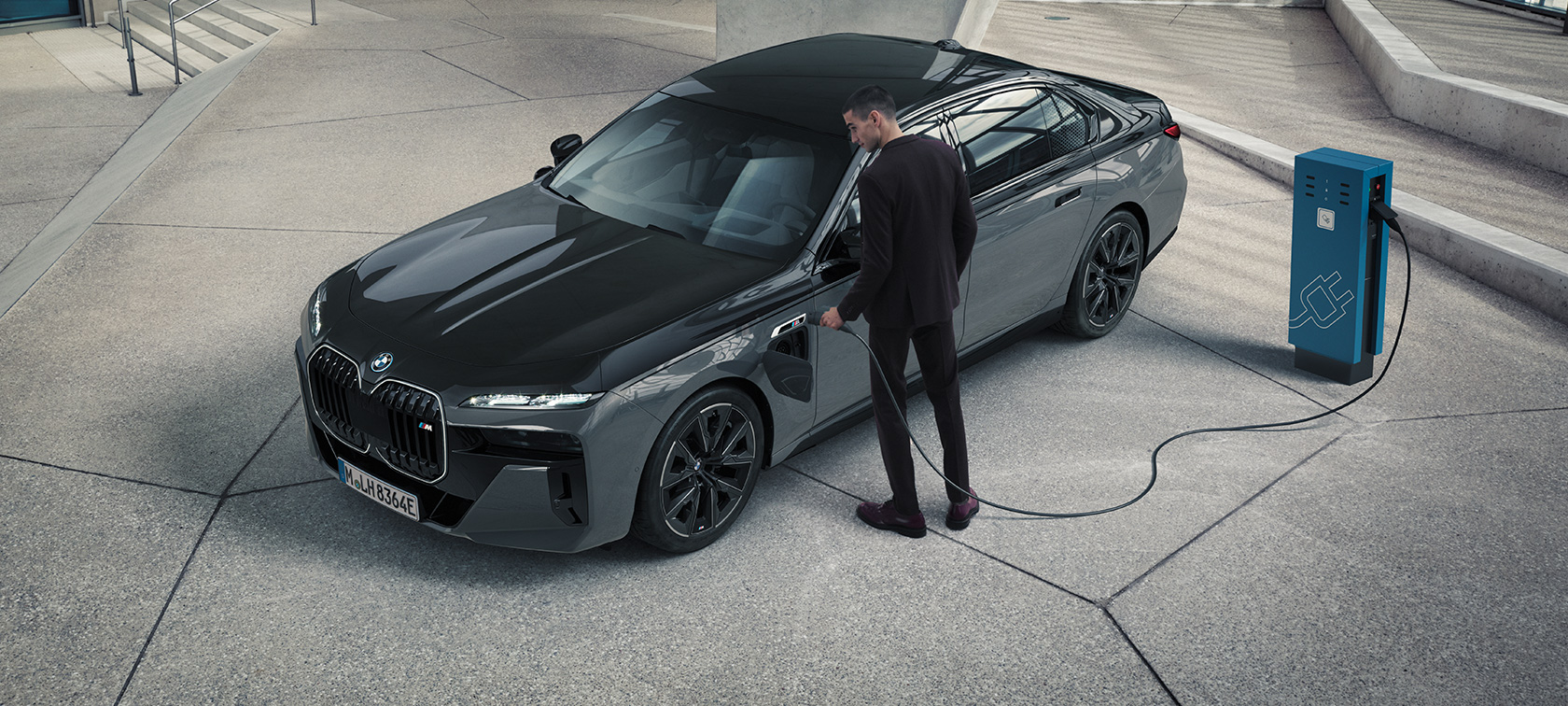 BMW M760e xDrive sedanas G70 M Performance paketas BMW Individual dviejų atspalvių Black Sapphire juoda metalizuota dažų apdaila BMW Individual Dravite Grey pilka metalizuota, trijų ketvirčių vaizdas iš priekio, vyras su įkrovimo laidu šalia įkrovimo stotelės