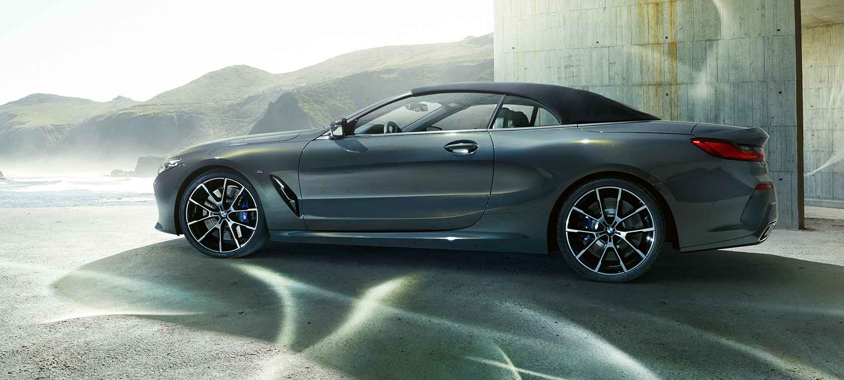 BMW M850i xDrive kabrioletas, vaizdas iš šono. BMW M850i xDrive kabrioletas Dravit Grey pilko metaliko spalvos su uždaru minkštu stogu, šoninis vaizdas kalvų fone.
