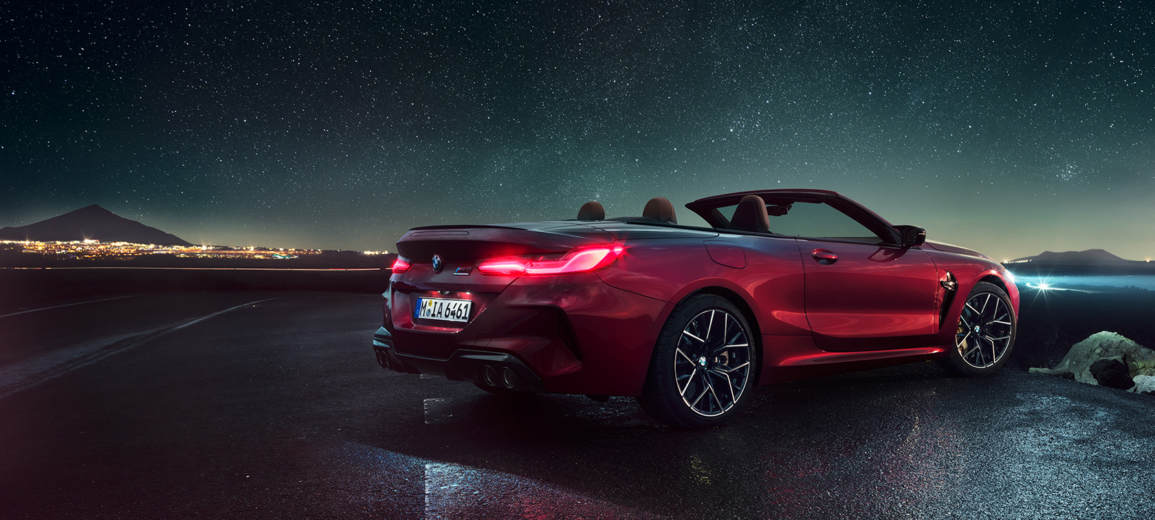 BMW M8 Competition kabrioletas, trijų ketvirčių vaizdas iš galo. BMW M8 Competition kabrioletas Motegi Red raudono metaliko spalvos su atviru minkštu stogu, trijų ketvirčių žvaigždėto dangaus fone stovinčio automobilio vaizdas iš galo.