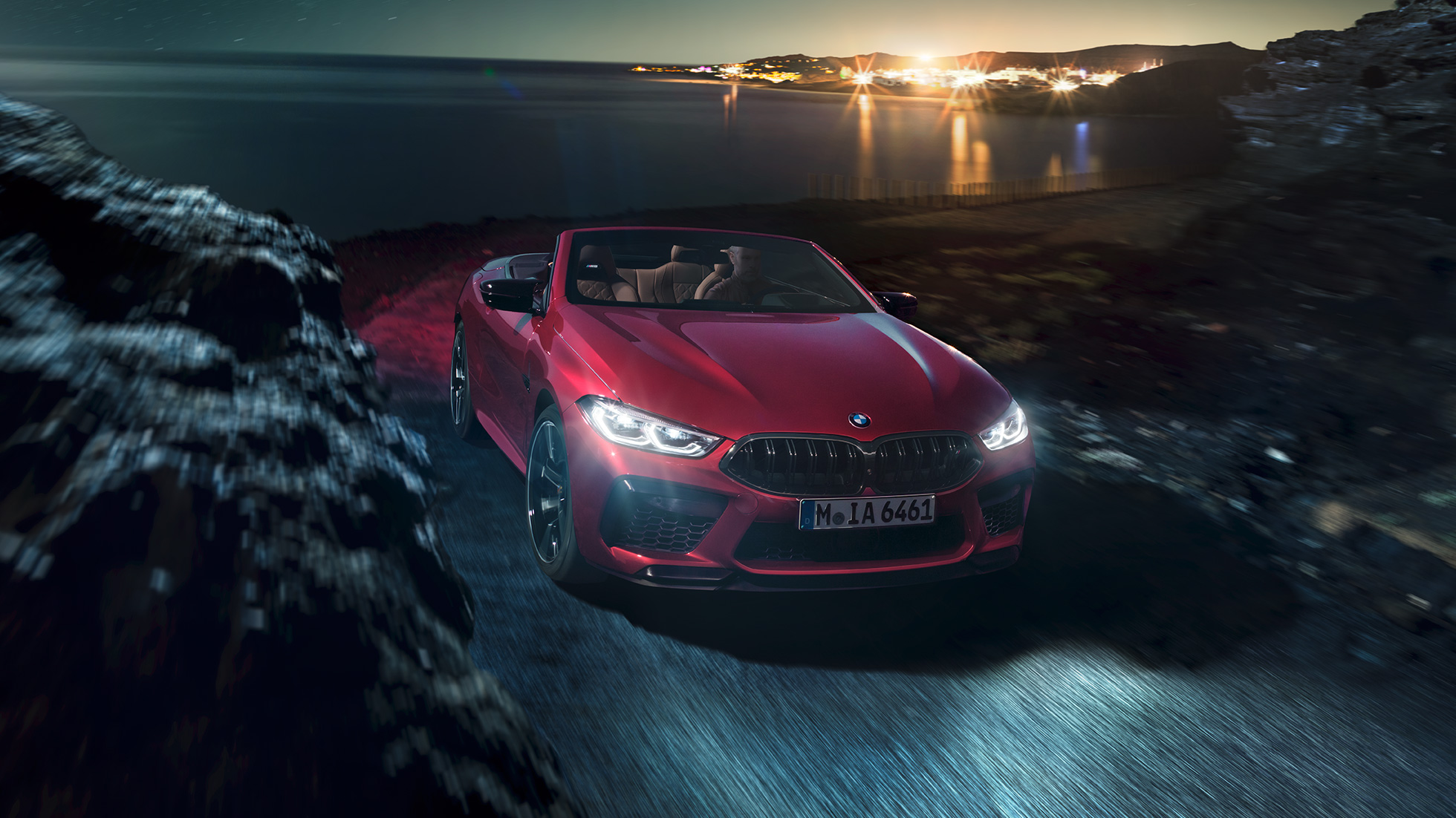 BMW M8 Competition kabrioletas, trijų ketvirčių vaizdas iš priekio. BMW M8 Competition kabrioletas Motegi Red raudono metaliko spalvos, su atviru minkštu stogu trijų ketvirčių naktinis važiuojančio pakrante automobilio vaizdas iš priekio.