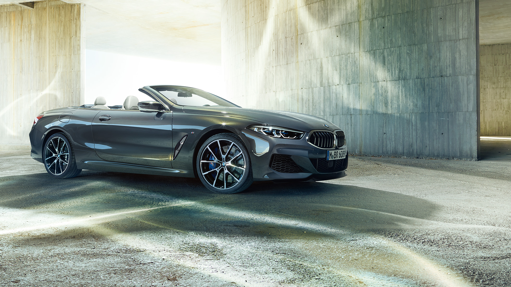 BMW M850i xDrive kabrioletas, trijų ketvirčių vaizdas iš šono. BMW M850i xDrive kabrioletas Dravit Grey pilko metaliko spalvos su atviru stogu, trijų ketvirčių priešais pastatą stovinčio automobilio vaizdas iš šono.