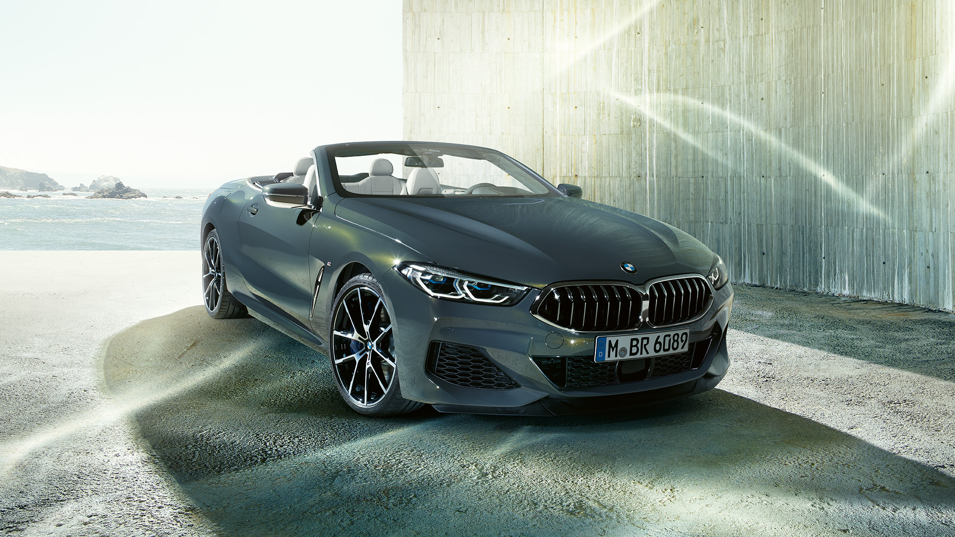 BMW M850i xDrive kabrioletas, trijų ketvirčių vaizdas iš priekio. BMW M850i xDrive kabrioletas Dravit Grey pilko metaliko spalvos su atviru minkštu stogu, trijų ketvirčių pakrantės fone stovinčio automobilio vaizdas iš priekio.