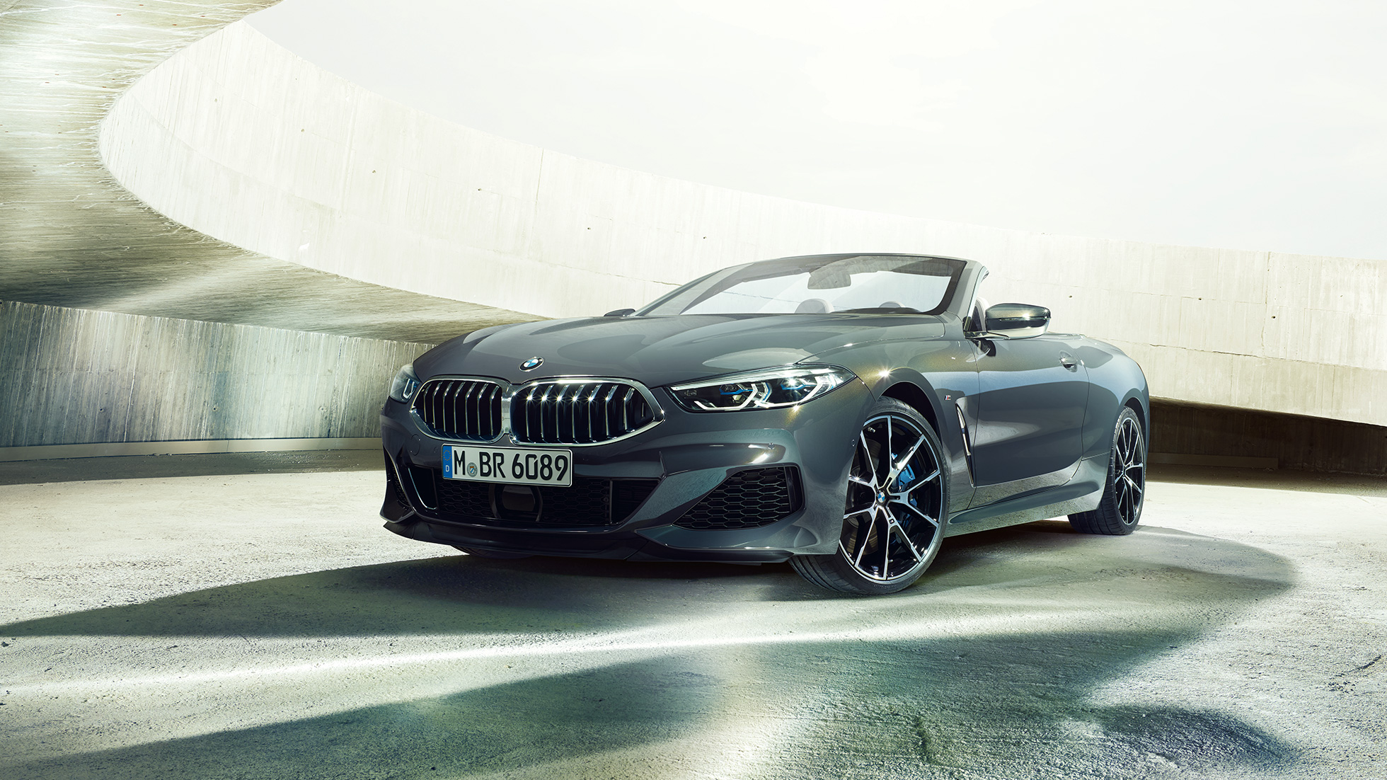 BMW M850i xDrive kabrioletas, trijų ketvirčių vaizdas iš priekio. BMW M850i xDrive kabrioletas Dravit Grey pilko metaliko spalvos, trijų ketvirčių vaizdas iš priekio, stovintis priešais betoninį pastatą.