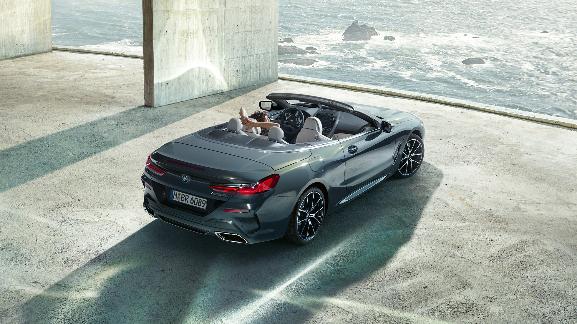 BMW M850i xDrive kabrioletas, vaizdas iš paukščio skrydžio. BMW M850i xDrive kabrioletas Dravit Grey pilko metaliko spalvos su atviru minkštu stogu, vaizdas iš paukščio skrydžio, automobilis parkuojamas pastate vandenyno fone.