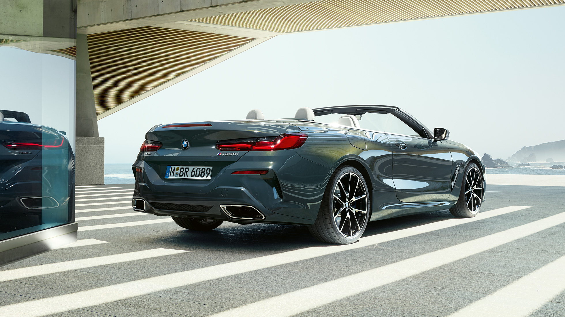 BMW M850i xDrive kabrioletas, trijų ketvirčių vaizdas iš galo. BMW M850i xDrive kabrioletas Dravit Grey pilko metaliko spalvos su atviru minkštu stogu, trijų ketvirčių parkuojamo automobilio vaizdas iš galo vandenyno fone.