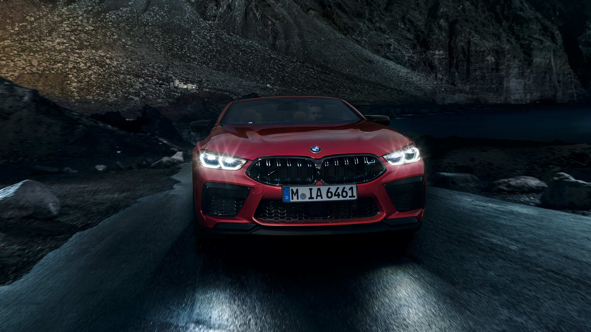 BMW M8 Competition kabrioletas, vaizdas iš priekio. BMW M8 Competition kabrioletas Motegi Red raudono metaliko spalvos, su šviečiančiais priekiniais žibintais, važiuojančio naktį automobilio vaizdas iš priekio.