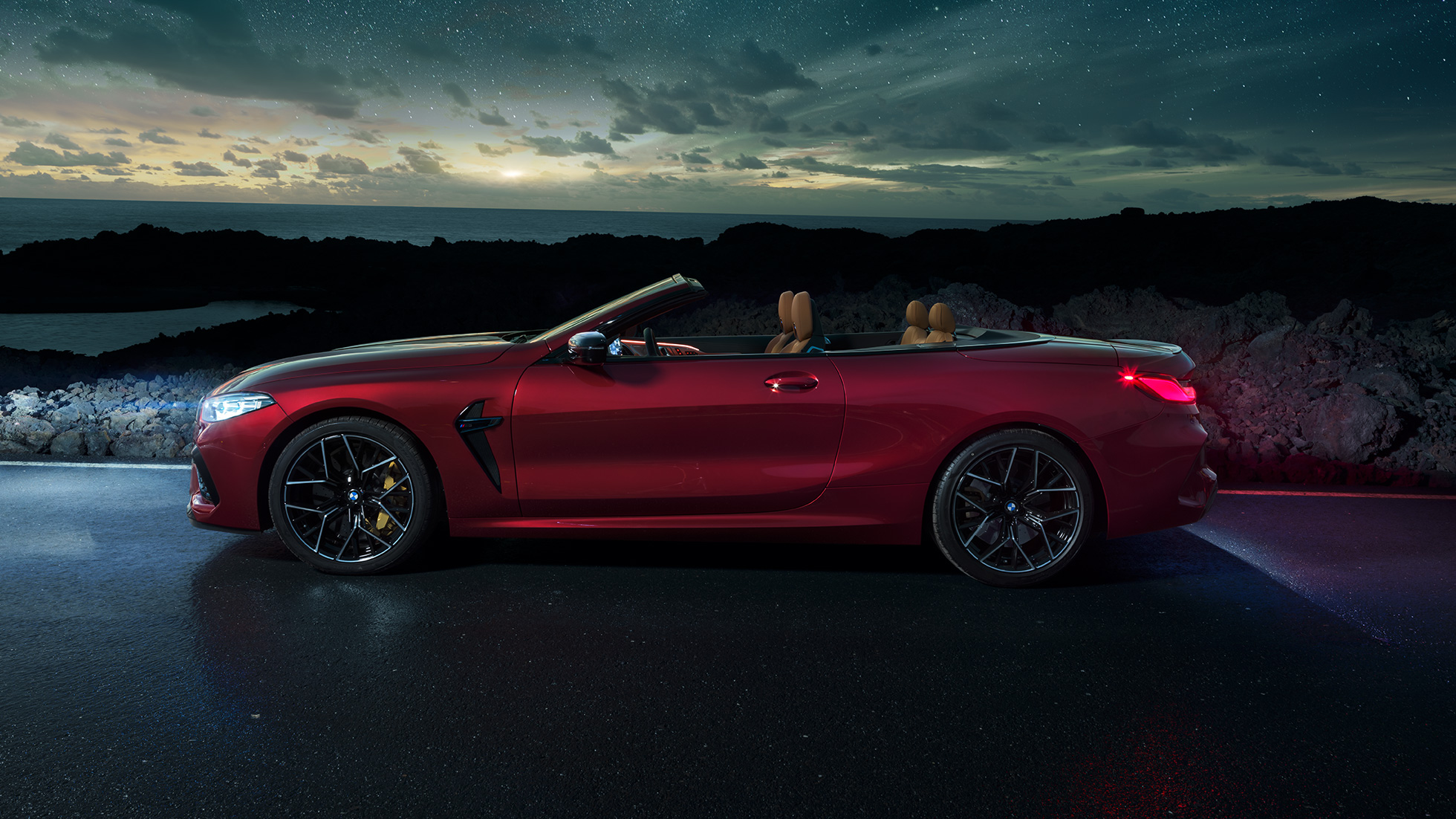 BMW M8 Competition kabrioletas, vaizdas iš šono. BMW M8 Competition kabrioletas Motegi Red raudono metaliko spalvos su atviru minkštu stogu, automobilio, stovinčio šalia vyro žvaigždėto dangaus fone, vaizdas iš šono.
